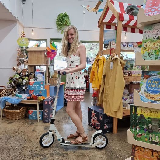 bree owner earth toys in cairns shop standing on her white mirco scooter classic adults scooter surrounded by colourful toys on wooden shelves, books and games