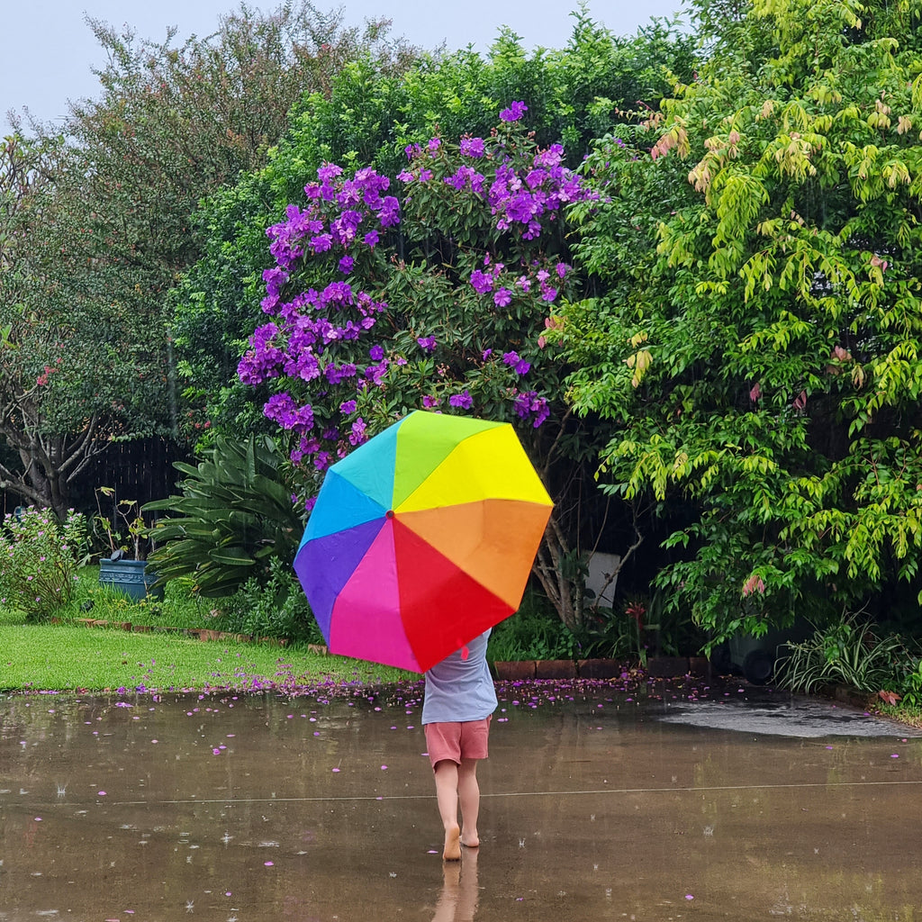 young boy holding compact rainbow large umbrella in the rain with trees and flowers